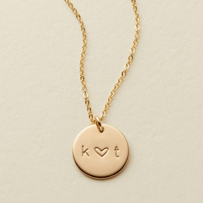 A gif of a delicate gold pendant chain necklace with a round disc charm, with the initials 'k' and 't' engraved. The symbol between them switches between '+', '&', and a heart. 