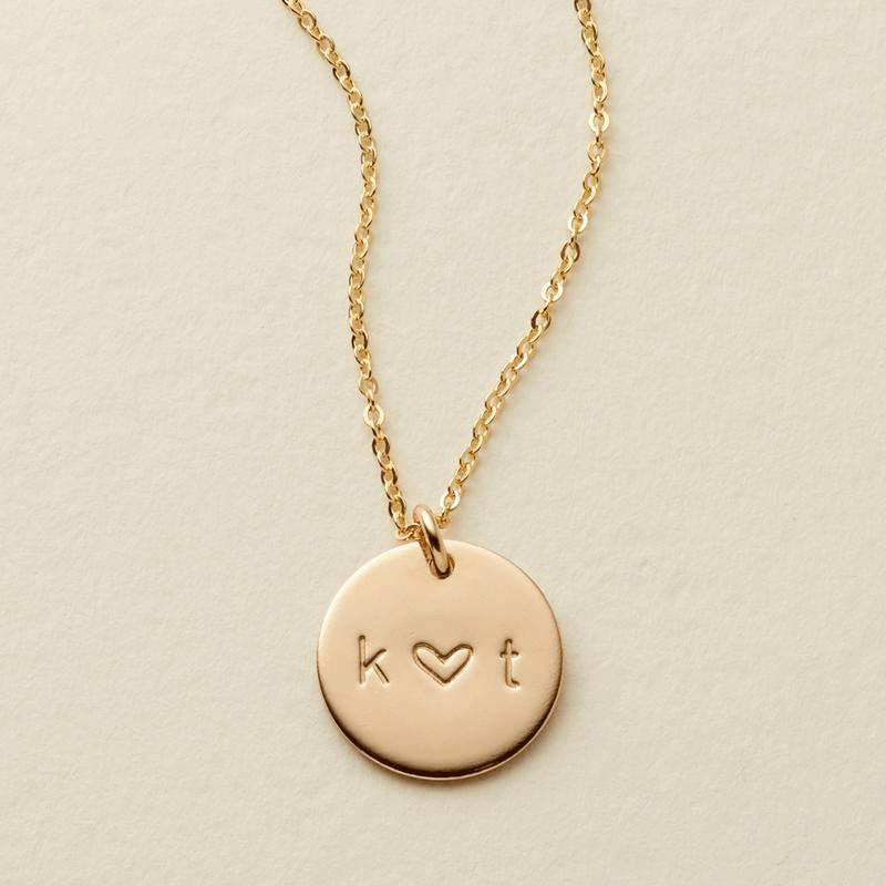 A gif of a delicate gold pendant chain necklace with a round disc charm, with the initials 'k' and 't' engraved. The symbol between them switches between '+', '&', and a heart. 