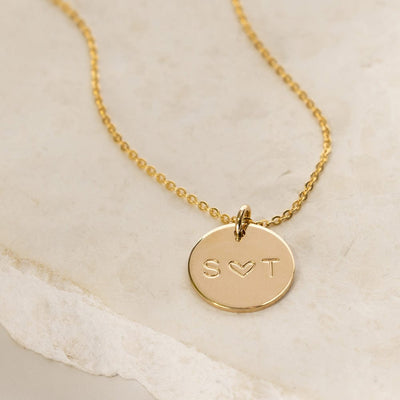 A gold pendant necklace with a round flat disc charm engraved with 'S ♥ T', shown on a light beige flat rock surface.