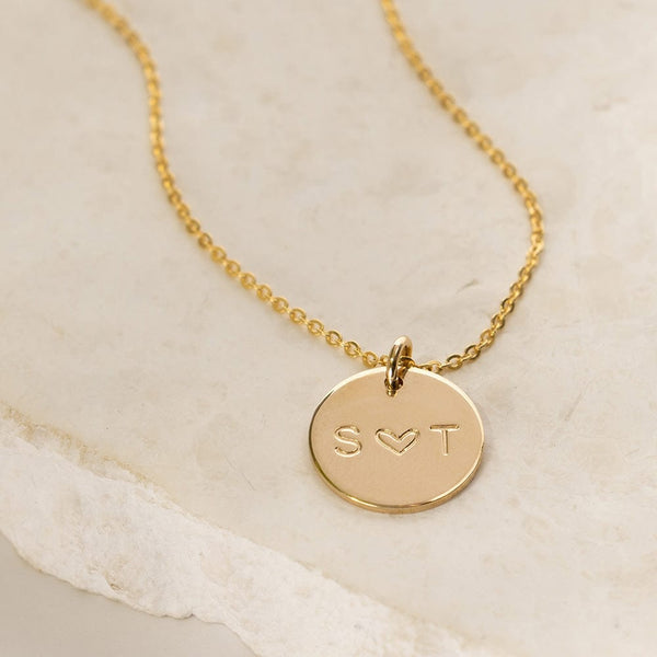 A gold pendant necklace with a round flat disc charm engraved with 'S ♥ T', shown on a light beige flat rock surface.