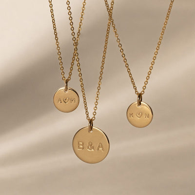 Three delicate gold pendant chain necklaces with engraved round disc charms, shown in front of an off-white background with shadows. Each disc is engraved with either 'A ♥ M', 'K♥ N' or 'B & A'.