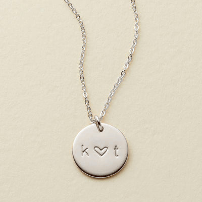 A delicate silver pendant chain necklace with an engraved disc reading 'k ♥ t', shown against a solid light beige background.