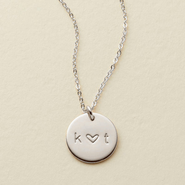 A delicate silver pendant chain necklace with an engraved disc reading 'k ♥ t', shown against a solid light beige background.