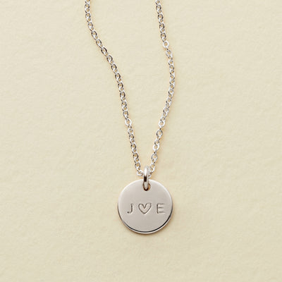 A delicate silver pendant chain with an engraved disc reading 'J ♥ E', shown on a solid light beige background.