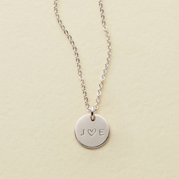 A delicate silver pendant chain with an engraved disc reading 'J ♥ E', shown on a solid light beige background.