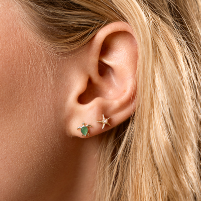 Close-up of an ear wearing two stud earrings, one with a turtle design and green gemstone and the other a star fish design with a pearl in the center.
