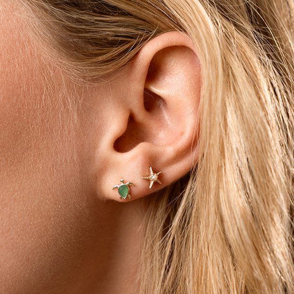 Close-up of an ear wearing two stud earrings, one with a turtle design and green gemstone and the other a star fish design with a pearl in the center.