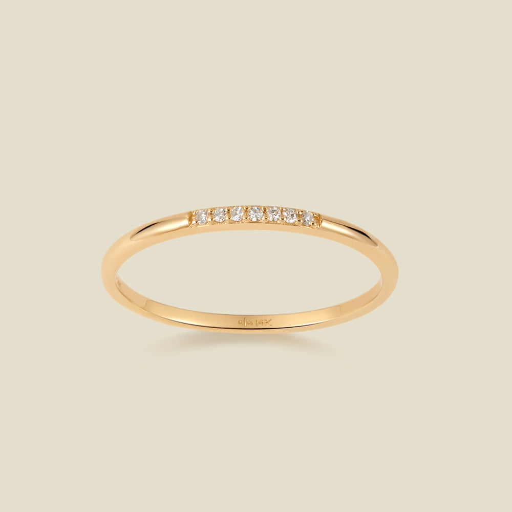 A slim yellow gold band ring featuring a row of small clear pavé-set diamonds across the front, shown on a light beige background. 