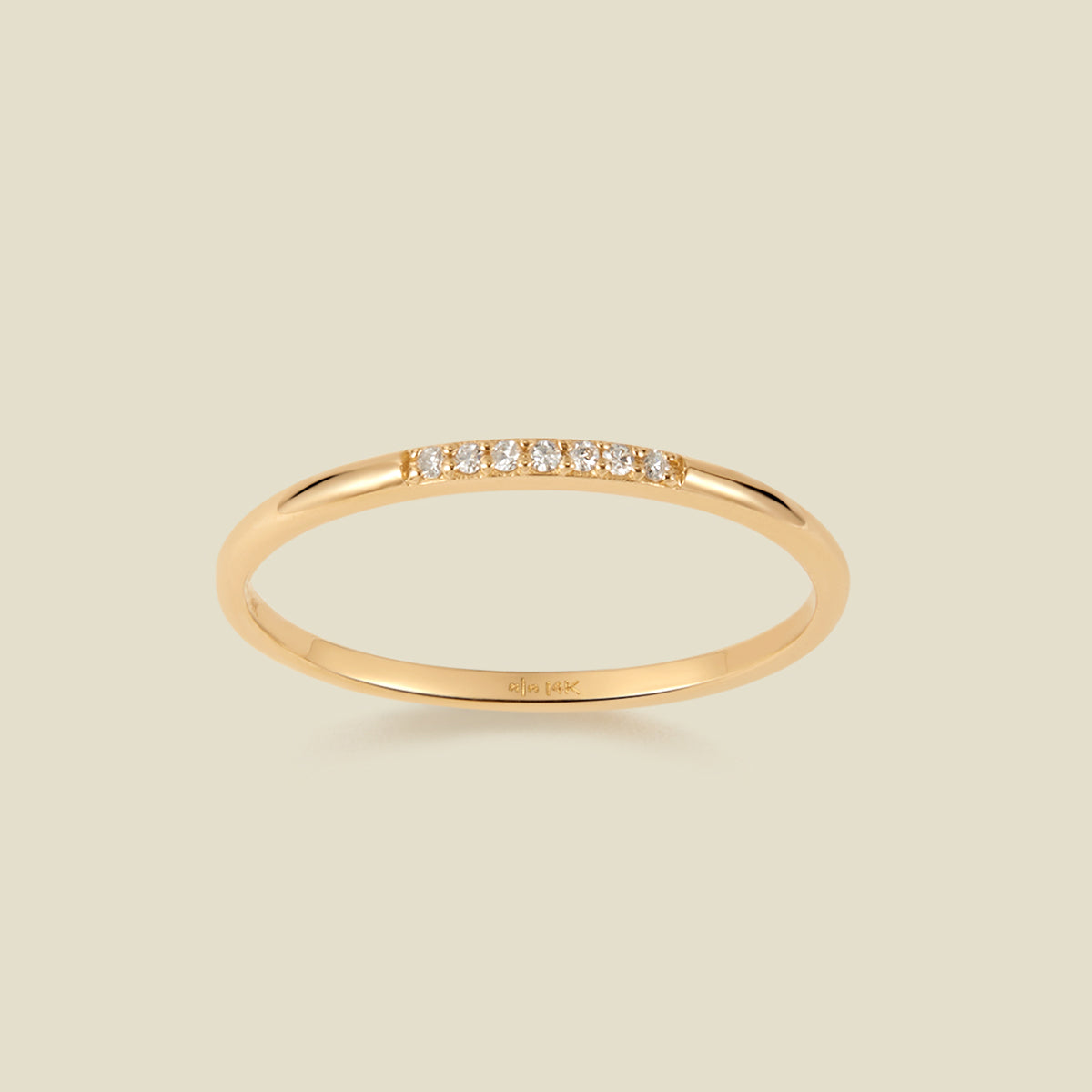 A slim yellow gold band ring featuring a row of small clear pavé-set diamonds across the front, shown on a light beige background. 