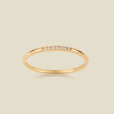 A slim yellow gold band ring featuring a row of small clear pavé-set diamonds across the front, shown on a light beige background. 