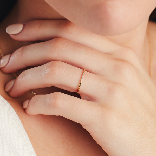 A close-up of a hand resting under a person's chin, showcasing a thin gold band ring with a single row of small circular diamonds across the front.