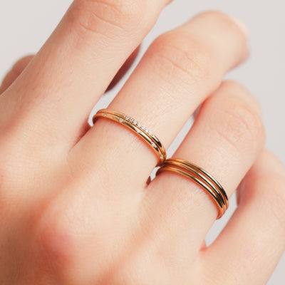 A close-up of a persons hand wearing three thin stacked gold rings on one finger and two stacked gold rings, each with a different pattern of clear gemstones, on another.