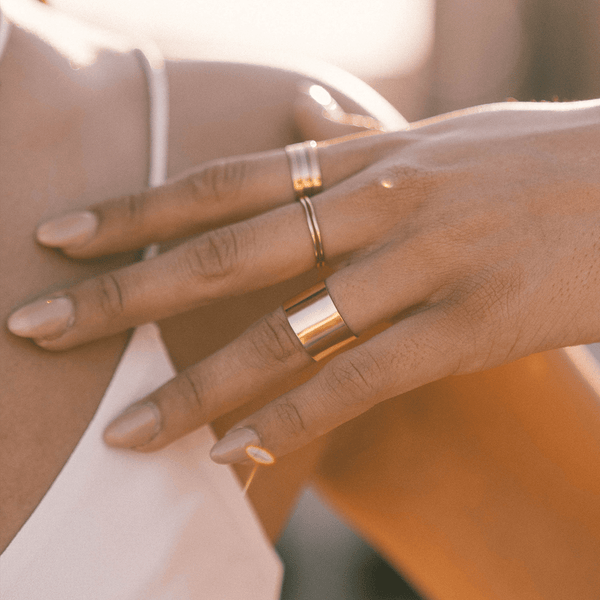 A person's hand wearing multiple gold rings, including two stacked thin wire rings and a large wide cigar band cuff ring.