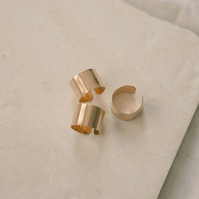 Three wide polished gold cigar band rings sitting in different positions on a textured off-white background.