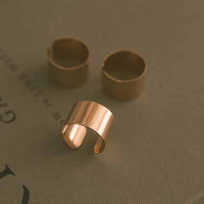 Three polished gold wide cigar band rings, showing an opening on the back and sitting on a textured grey book.