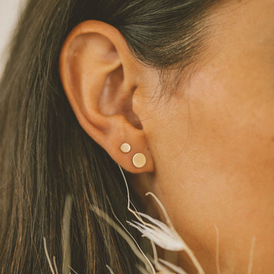 A person's ear wearing two pairs of gold round flat disc studs in two sizes.