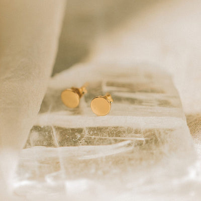 A pair of flat round gold disc stud earrings sitting on a clear, jagged textured rock.