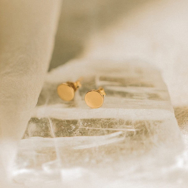 A pair of flat round gold disc stud earrings sitting on a clear, jagged textured rock.