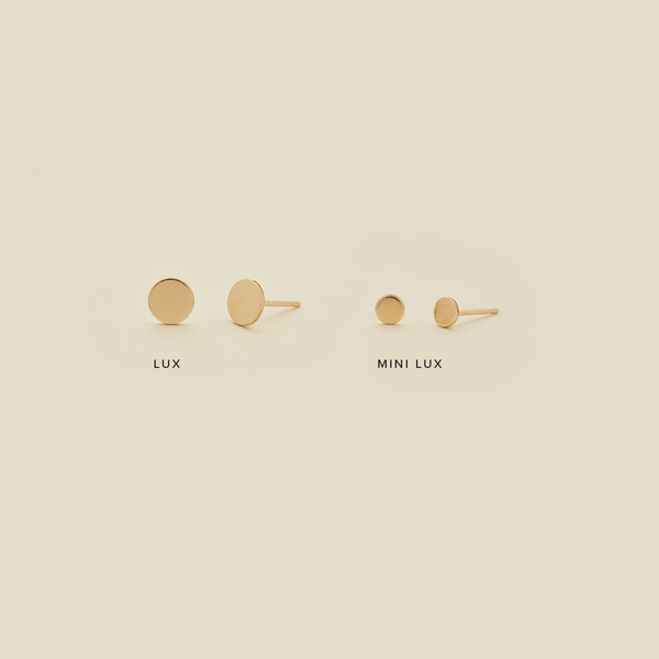 Two pairs of round gold flat disc stud earrings in different sizes, shown on a solid light beige background. Each has text underneath, with the larger stud saying 'LUX' and the smaller earrings saying 'MINI LUX'.