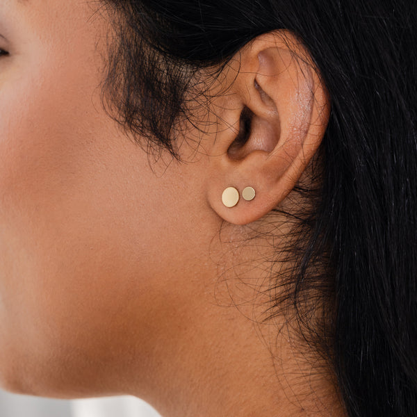 A close-up of a person's ear wearing two pairs of flat round gold disc studs in different sizes.