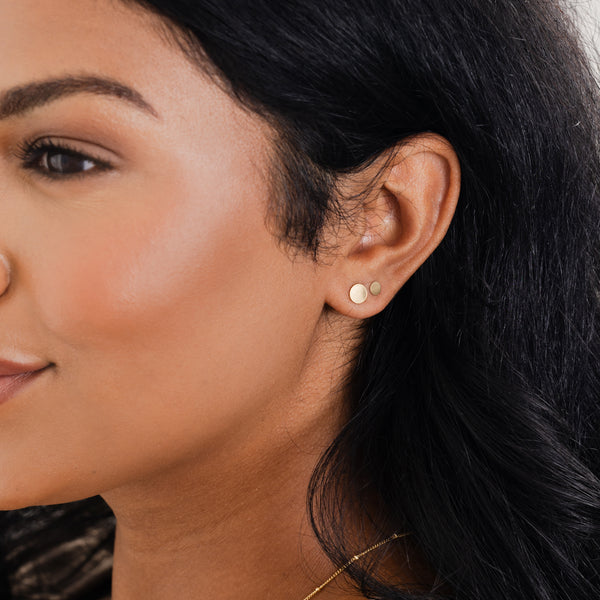 A close-up of a person's ear wearing two pairs of flat round gold disc studs in different sizes.