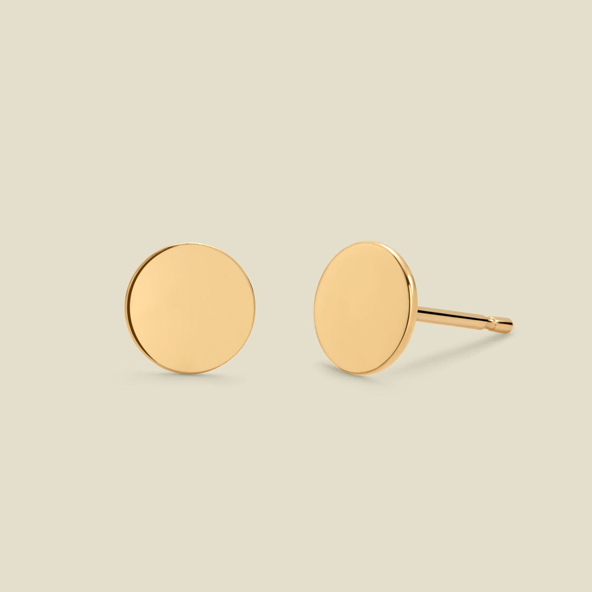 A pair of minimalist round gold disc stud earrings shown from two angles (front and side) on a solid light beige background.