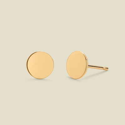 A pair of minimalist round gold disc stud earrings shown from two angles (front and side) on a solid light beige background.