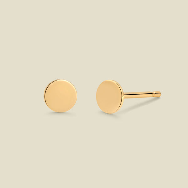 A pair of minimalist round gold disc stud earrings shown from two angles (front and side) on a solid light beige background.