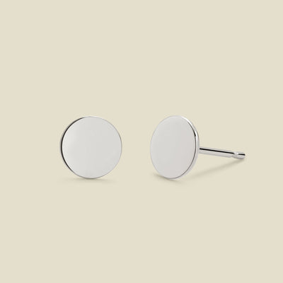 A pair of minimalist round silver disc stud earrings shown from two angles (front and side) on a solid light beige background.