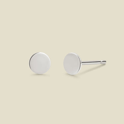 A pair of minimalist round silver disc stud earrings shown from two angles (front and side) on a solid light beige background.