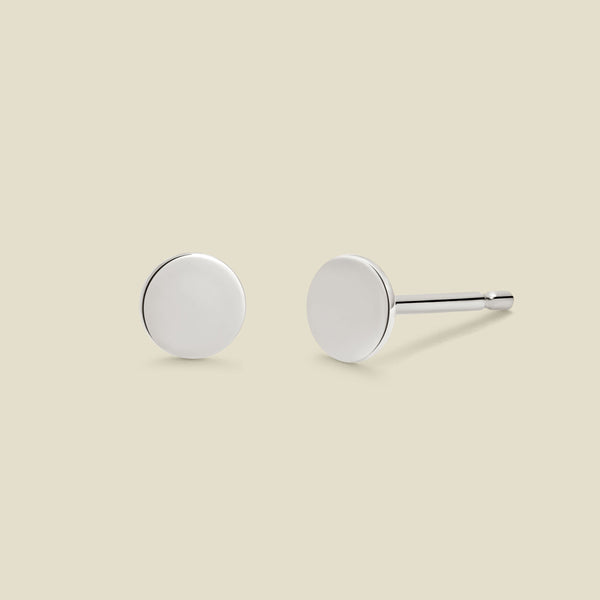 A pair of minimalist round silver disc stud earrings shown from two angles (front and side) on a solid light beige background.