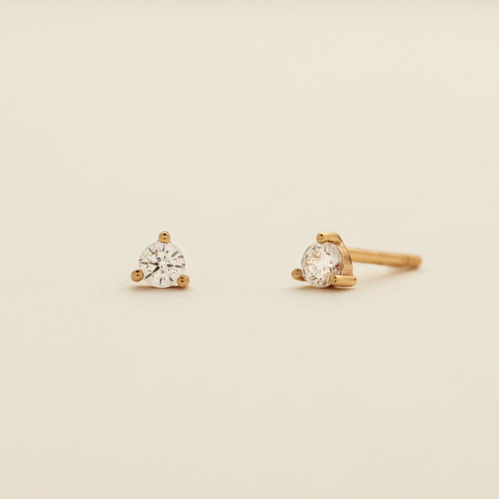 A pair of gold stud earrings with clear prong-set gemstones, shown on a plain light beige background.