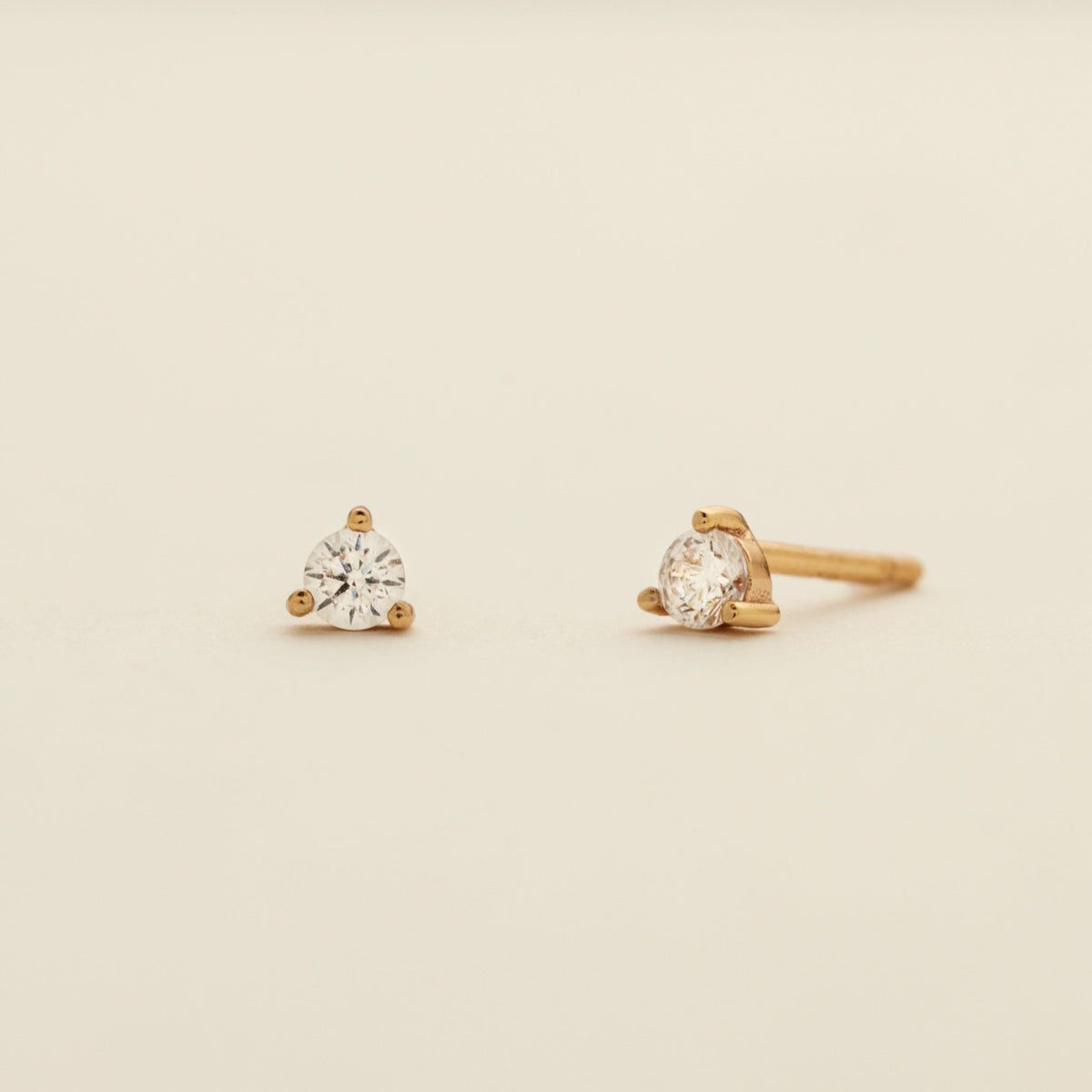 A pair of gold stud earrings with clear prong-set gemstones, shown on a plain light beige background.