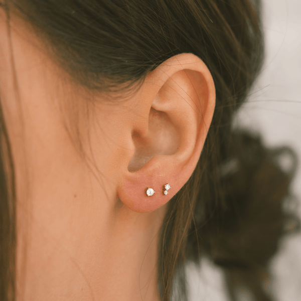 A woman's ear wearing a gold stud with a prong-set clear gemstone and a stud with two clear gemstones set vertically with the top being larger than the bottom.