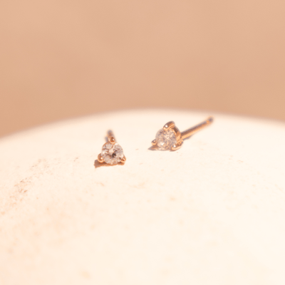 A pair of gold stud earrings with a prong-set clear gemstone, shown on a textured light beige surface with a spotlight of light on them.