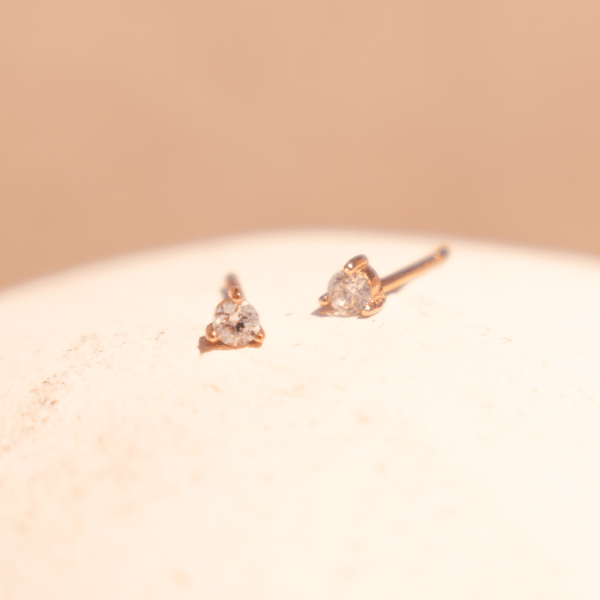 A pair of gold stud earrings with a prong-set clear gemstone, shown on a textured light beige surface with a spotlight of light on them.