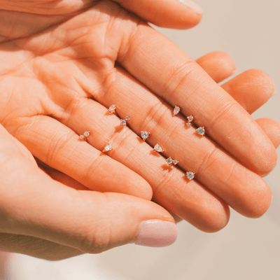 A person's hand with multiple styles of gold stud earrings sitting between their fingers, each with different patterns and shapes of clear gemstones.