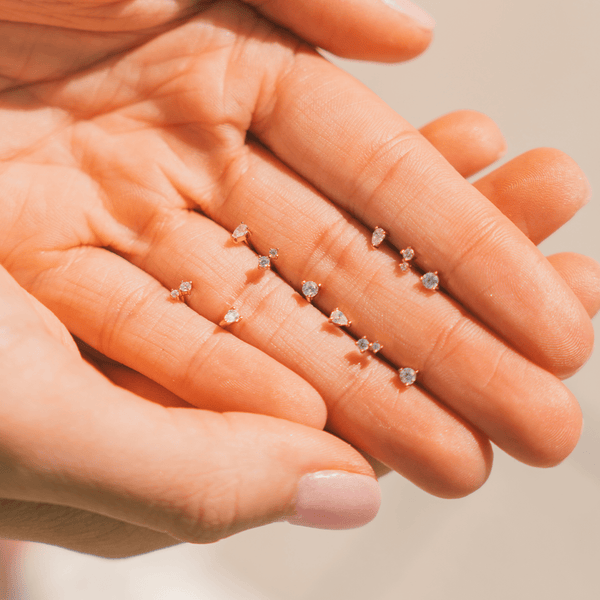 A person's hand with multiple styles of gold stud earrings sitting between their fingers, each with different patterns and shapes of clear gemstones.