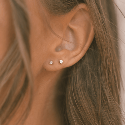 A close-up of a person's ear wearing two gold stud earrings, each with a prong-set clear gemstone. One is round, and the other is a teardrop shape.