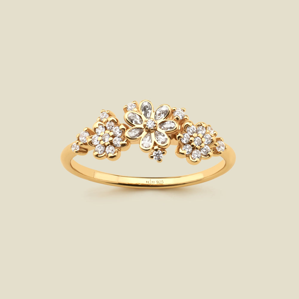 A yellow gold ring featuring three stylized flower clusters set with clear stones, photographed against a solid light beige background. 