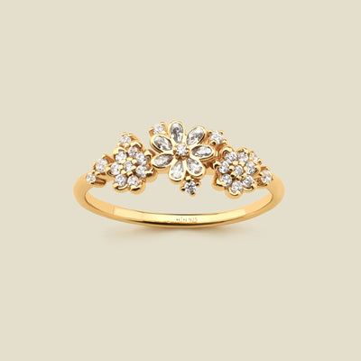 A yellow gold ring featuring three stylized flower clusters set with clear stones, photographed against a solid light beige background. 