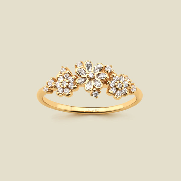 A yellow gold ring featuring three stylized flower clusters set with clear stones, photographed against a solid light beige background. 