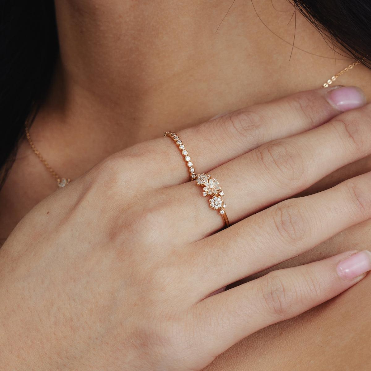 A person's hand wearing a thin gold band ring made up of small round bezel-set clear gemstones, and a bold gold ring with floral clusters made of clear gemstones.