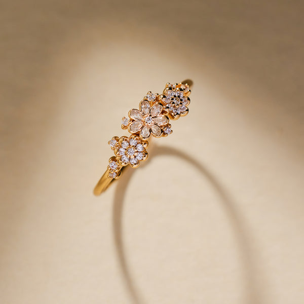 A yellow gold ring featuring three stylized flower clusters set with clear stones, photographed against a shadowed light beige background. 