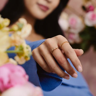 A person's hand wearing a thin gold band ring made up of small round bezel-set clear gemstones, and a bold gold ring with floral clusters made of clear gemstones.