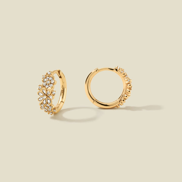 A pair of gold huggie hoop earrings on a solid light beige background. One earring is shown from the front, featuring a cluster of clear floral-cut stones, while the other is shown from the side showing a textured edge.
