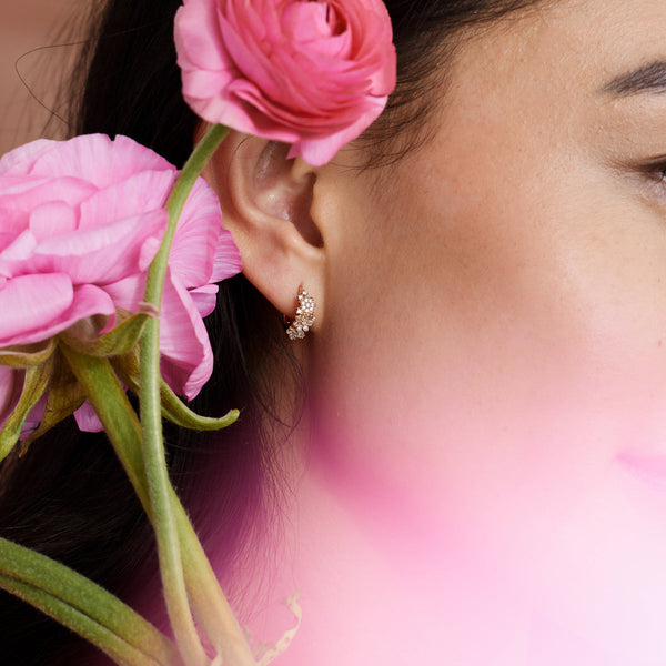 A close-up of a person's ear wearing a gold huggie hoop earring with clear gemstone flowers covering the front, with pink and red flowers in the foreground.