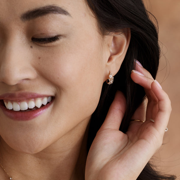 A close-up of a person's ear wearing a gold huggie hoop covered in clear gemstones making up flower clusters, paired with gold rings and set against a warm light brown background.
