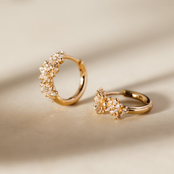 A pair of gold huggie hoop earrings with clusters of clear stones creating a flower design, set against a light beige background with shadows.