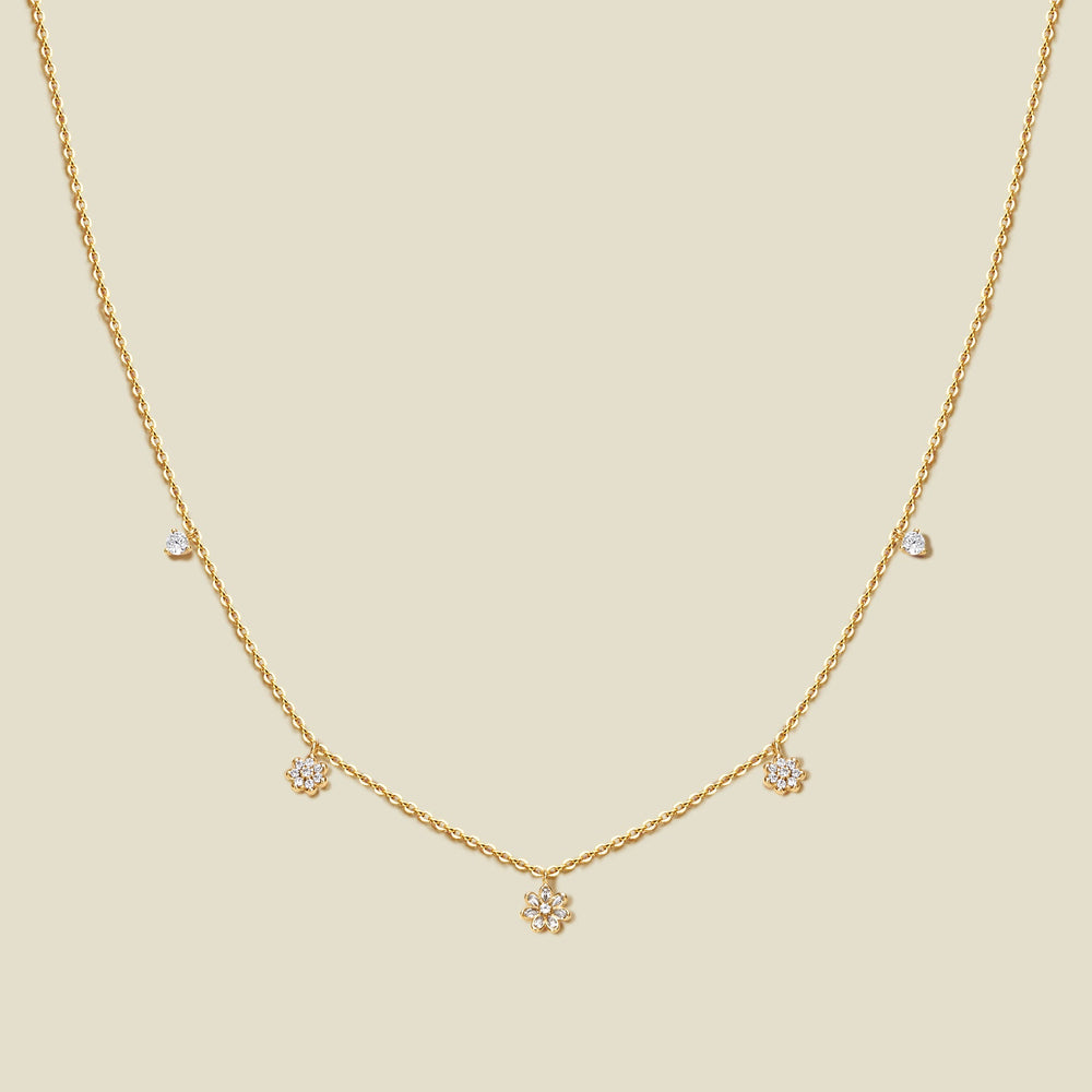 A delicate gold chain necklace with three small flowers with clear accent stones and a few tiny clear accent stones, displayed on a solid light beige background.
